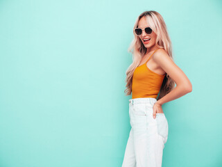Fototapeta na wymiar Young beautiful smiling blond female in trendy summer yellow clothes. Sexy carefree woman posing near blue wall in studio. Positive model having fun indoors. Cheerful and happy. In sunglasses
