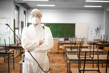Fototapeta na wymiar Portrait of man in white sterile protection suit disinfecting and sanitizing desks and chairs in school classroom.