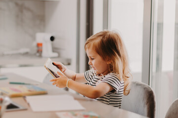 Cute toddler girl wear casual t-shirt with stripes sitting in kitchen with smartphone.