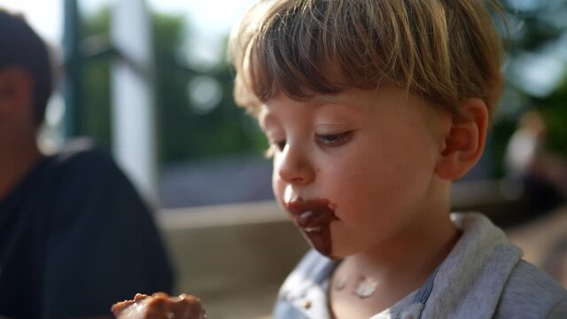 Messy baby eating chocolate ice-cream cone outside, cute toddler boy covered with icream