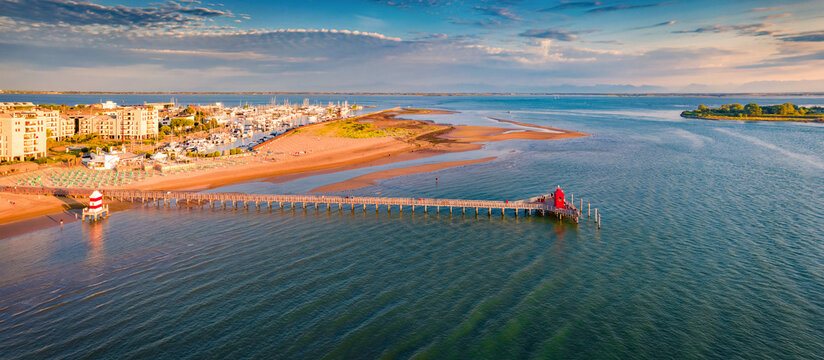 Panoramaic summer view from flying drone of Lignano Sabbiadoro town. Sunny outdoor scene of Adriatic coast of Italy with lighthous and old wooden pier. Amazing morning seascape of Mediterranean sea.