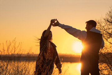 Young romantic couple in casual style dancing at sunset by pond. Sun reflected in water. Back view. Youth, love and freedom concept. Soft focus. Warm toned.