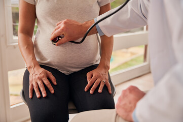 Caucasian pregnant female sitting in clinic while male doctor examines pregnant belly with...