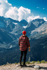 Fototapeta na wymiar A traveler on the background of mountains. A tourist stands on top of a mountain. Male traveler in the mountains, rear view. A traveler in a red plaid shirt and a red hat. Domestic tourism. Copy space