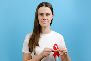 Young brunette woman 20s holds red ribbon, looking at camera, isolated on blue color background studio. Blood transfusion and donation. Hemophilia and World Health concept. World AIDS and hiv day - obrazy, fototapety, plakaty