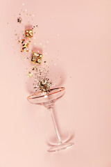 Modern champagne glass with splash of confetti and decorations over pink background.