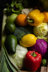 Ripe colorful vegetables and fruits: bell pepper, cabbage, avocado, leek, orange, lemon in a wooden box, black background. Close view.