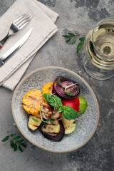 A stone plate with grilled vegetables on a gray table. Fried zucchini, eggplant, corn and grilled mushrooms, top view