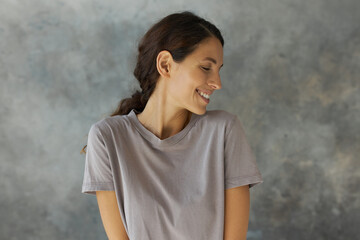 Profile close-up portrait of charismatic brown-haired Caucasian woman with charming smile and closed eyes, looking aside with dreamy face expression, posing against grey background