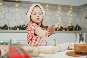 Little girl 3 years old in red Christmas cap and checkered shirt prepares dough for gingerbread cookies in kitchen