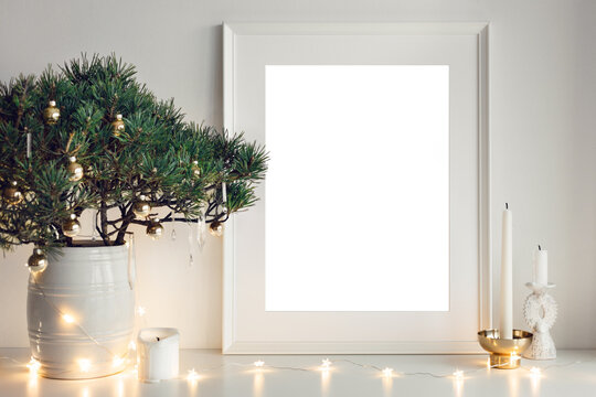 Mockup template. Christmas room decor, white medium size photo frame with blank copy space for your advertising content, candles and decorated coniferous bonsai. Festive holidays preparation