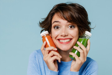 Young happy woman 20s in casual sweater hold pressed juice green orange vegetable smoothie as detox diet isolated on plain pastel light blue background studio portrait. People lifestyle food concept.