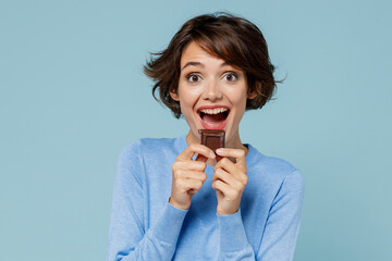 Young smiling happy woman in casual sweater hold sweet pink cream donuts biting chocolate bar look camera isolated on plain pastel light blue background studio portrait. People lifestyle food concept.