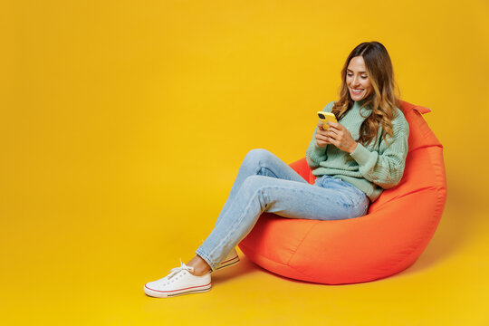 Full Body Young Smiling Woman 30s In Green Knitted Sweater Sit In Bag Chair Hold Use Mobile Cell Phone Chatting Browsing Internet Isolated On Plain Yellow Background Studio. People Lifestyle Concept.