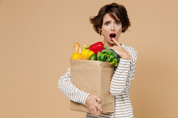 Young shocked scared astonished surprised vegetarian woman in casual clothes hold paper bag with vegetables look camera with opened mouth isolated on plain pastel beige background Shopping concept.