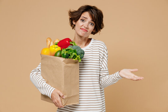 Young uncertain vegetarian woman wear casual clothes hold paper bag with vegetables after shopping in greengrocery shrugging shoulders spread hand isolated on plain pastel beige background studio.