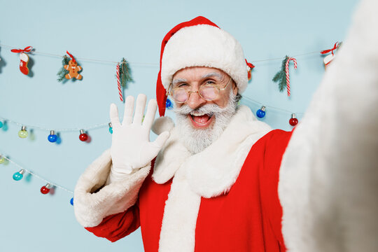 Close up old bearded Santa Claus man 50s in Christmas hat red suit do selfie shot pov on mobile phone waving hand isolated on plain blue background studio Happy New Year 2022 merry ho x-mas concept.
