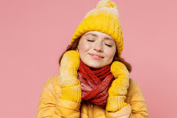 Fascinating happy calm young woman 20s years old wears yellow jacket hat mittens keep eyes closed put hands on scarf wrapped around neck isolated on plain pastel light pink background studio portrait.