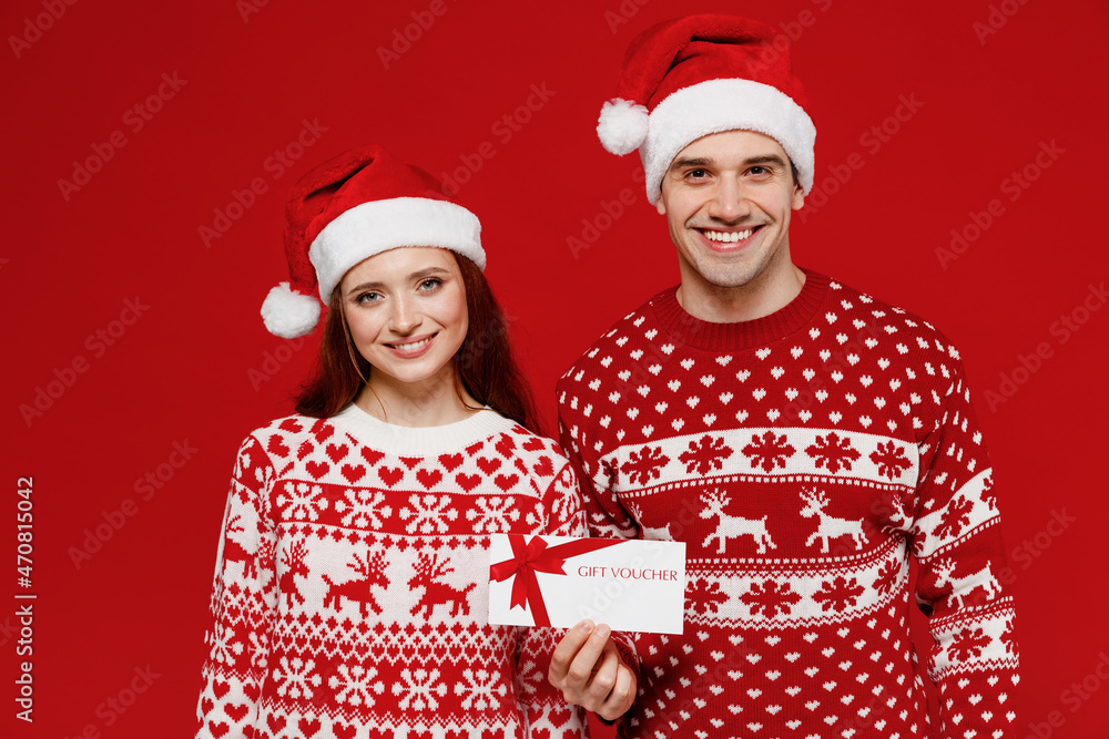 Wall mural Young couple friends two man woman in sweater hat hold gift certificate coupon voucher card for store isolated on plain red background. Happy New Year 2022 celebration merry ho x-mas holiday concept.