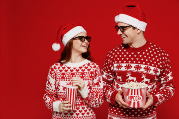 Young fun couple friend man woman in 3d glasses watch movie film hold bucket of popcorn cup of soda pop look to each other isolated on plain red background People emotions in cinema lifestyle concept