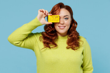 Young cheerful chubby overweight plus size big fat fit woman wear green sweater hold in hand cover eye with credit bank card isolated on plain blue background studio portrait People lifestyle concept.