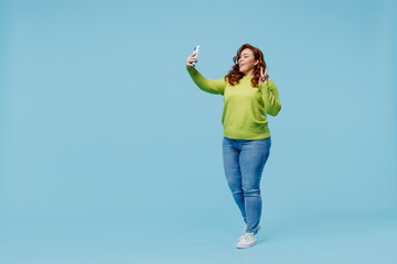Full body smiling young chubby overweight plus size big fat fit woman wear green sweater doing selfie shot on mobile cell phone show v-sign isolated on plain blue background People lifestyle concept.