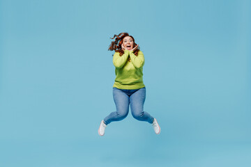 Young overjoyed fun excited surprised chubby overweight plus size big fat fit woman wear green sweater jump high hold face isolated on plain blue background studio portrait. People lifestyle concept