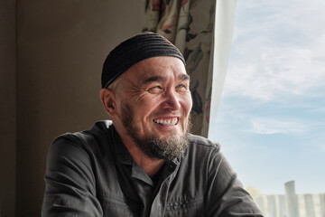 Portrait of happy senior asian muslim man with beard wearing skullcap sitting by window and smiling.