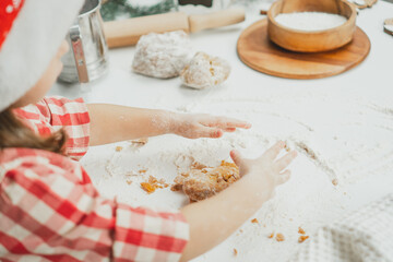 Partially blurred hands of little girl in Christmas cap and checkered shirt knit dough for cookies on white table