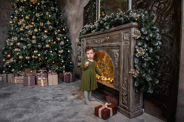 Little girl in a warm green dress holds a golden ball in hands, stands by a bright wooden fireplace near the Christmas tree with gifts. Decorating a stylish holiday apartment interior. Copy space