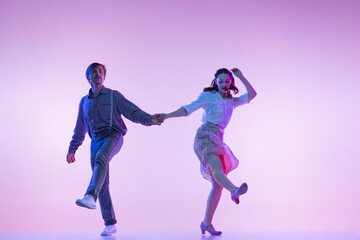 Obraz na płótnie Canvas Dynamic portrait of young dancers, man and woman in vintage style outfits dancing swing isolated on lilac color background in neon light