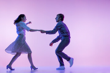 Two young graceful dancers, man and woman in vintage style outfits dancing rock-and-roll isolated on lilac color background in neon light