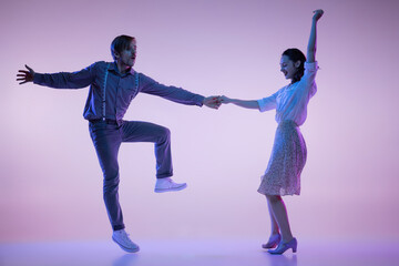 Fototapeta na wymiar Two young graceful dancers, man and woman in vintage style outfits dancing rock-and-roll isolated on lilac color background in neon light
