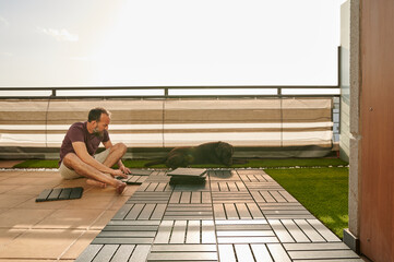 middle-aged man setting up an artificial floor on his terrace with his Labrador retriever laying next to him