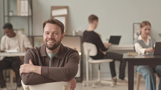 Locked-down of young Caucasian bearded man walking into frame, sitting back to front on chair in busy office on regular workday, looking and smiling on camera
