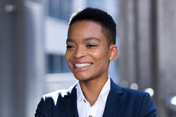 Headshot photo with close up portrait of young successful business woman, african american woman smiling and rejoicing looking at camera, in business suit, outside office