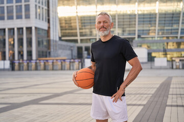 Active middle aged man in sportswear looking at camera, standing outdoors with basketball ball, ready for workout in the city