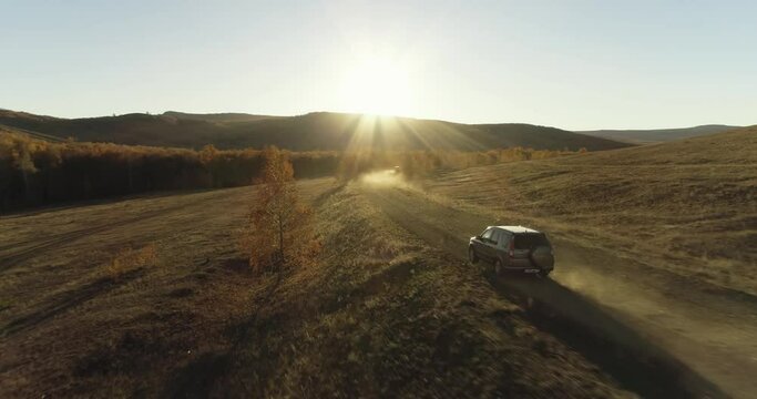 One gray crossover SUV car driving alone road trip, travel on gravel road through arid dry large field with vastness background at autumn sunny sunset - Aerial drone wide view