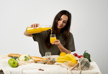 Woman in green t-shirt pouring a juice from a glass bottle in a glass over a table with mesh eco bag, healthy vegan vegetables, fruits, bread, snacks. Zero waste concept.
