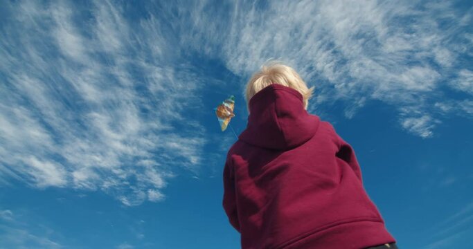 Rear view of happy baby boy child playing and flying kite outdoors at windy sunny day against the blue cloudy sky. Concept of carefree childhood kid playing with toy kite