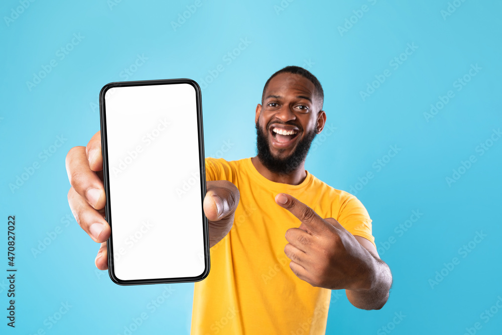 Poster Overjoyed black man pointing at cellphone with empty white screen on blue studio background, mockup for app or website