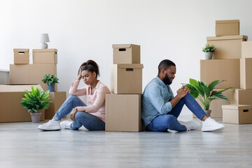 Unhappy young black guy and lady are tired and sitting on floor in apartment with boxes