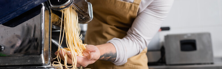 Cropped view of tattooed chef making spaghetti on pasta maker machine in kitchen, banner