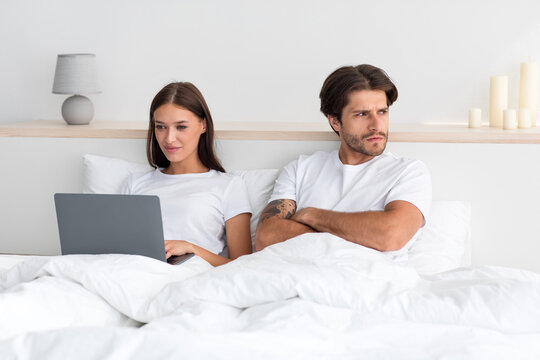 Young European Lady Work On Laptop In Bed, Ignoring To Disgruntled Man With Crossed Arms Looks To Side In Bedroom