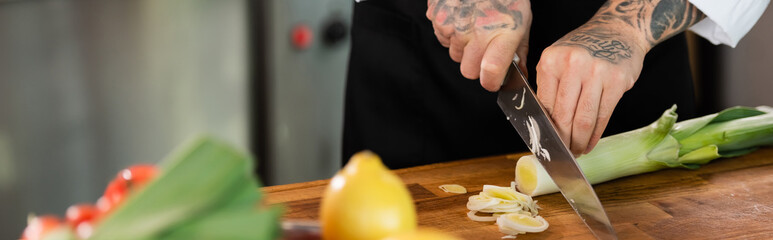 Cropped view of chef cutting leek near blurred vegetables in kitchen, banner