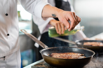 Cropped view of chef pouring salt while cooking meat on frying pan in kitchen