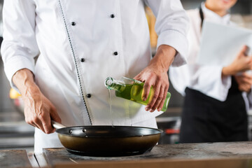 Cropped view of chef pouring olive oil on frying pan on cooktop in kitchen