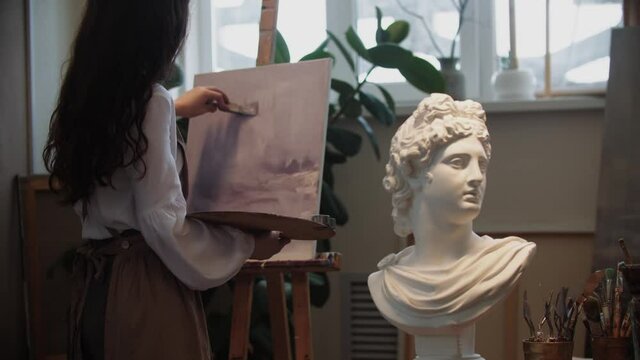Young woman artist with beautiful hair working with a painting - marble bust on the foreground