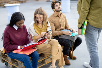 Multiracial students reading books while sitting on wooden bench outdoors at day. Concept of education and learning. Idea of student lifestyle. Young modern guys and girls at university campus