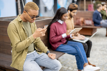 Multiracial students using gadgets outdoors. Concept of education. Remote and e-learning. Idea of students lifestyle. Young guys and girl sitting on wooden bench at university campus